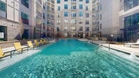 Located at 2124 American Industrial Way in Chamblee, Georgia, this expansive outdoor amenity space is centered around a resort-style swimming pool set within a striking courtyard environment. The pool itself is generously sized, featuring crystal-clear blue water, built-in water features including fountain jets and water wall elements, and a shallow sun ledge ideal for lounging. The pool deck is finished with clean, light-toned pavers and is lined with an extensive collection of chaise lounge chairs — both white and yellow-accented styles — providing ample space for sunbathing and relaxation. The courtyard is framed on multiple sides by a modern multi-story building with a contemporary façade combining brick and light stucco exteriors, large windows, and private balconies that overlook the amenity area. A distinctive steel truss architectural bridge element spans one end of the courtyard, adding an industrial-chic design detail that references the area's heritage. Additional outdoor seating areas with lounge sofas are nestled along the perimeter, accompanied by greenery and manicured landscaping. The space is enclosed by ornamental fencing while remaining open to natural light and sky views, creating an inviting and social outdoor retreat.