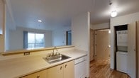 This single-floor residential unit offers a practical and comfortable layout designed for everyday living. The kitchen is positioned at the heart of the space, featuring light wood cabinetry, a stainless steel double-basin sink, white countertops, and a built-in dishwasher. The open design allows the kitchen to flow seamlessly into the living area, creating a connected and sociable environment. Natural light fills the living space through a large window that looks out onto a private balcony, offering pleasant outdoor views and enhancing the sense of openness throughout. The flooring transitions from the kitchen area into warm-toned hardwood in the adjacent spaces, adding a cohesive and inviting character to the interior. A hallway visible from the kitchen leads to additional rooms, including what appears to be a closet or storage space and further private areas of the unit. The overall aesthetic is clean and neutral, with white walls and recessed lighting contributing to a bright, well-lit ambiance. The layout is efficient, making thoughtful use of the available footprint while maintaining a comfortable sense of space throughout each area of the home.