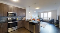 The Cobalt presents a thoughtfully designed single-floor residence that blends contemporary style with functional living. The open-concept layout connects the kitchen and living area seamlessly, creating a spacious and inviting atmosphere filled with natural light from large windows.

The kitchen is a standout feature, showcasing rich wood-tone cabinetry with sleek hardware, dark granite-style countertops, and a full suite of stainless steel appliances including a range, built-in microwave, and dishwasher. A mosaic tile backsplash adds a refined textural element, while pendant lighting suspended above the peninsula provides both task illumination and a modern aesthetic touch. The peninsula itself offers additional counter space and casual seating potential, making it ideal for entertaining or everyday dining.

Flowing directly from the kitchen, the living area is bright and welcoming, benefiting from generous window coverage that draws in ample natural light. Rich dark hardwood flooring runs throughout, anchoring the space with warmth and continuity. The overall ambiance is modern and polished, with clean lines and a neutral palette that offers a versatile backdrop for a variety of interior styles. The Cobalt delivers a well-appointed, move-in-ready living experience with attention to quality finishes throughout.