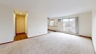 Located at 7635 Greenfield Ave in Mounds View, Minnesota, this two-bedroom, one-bathroom unit offers a well-organized single-floor layout designed for comfortable everyday living. The open living and dining area is generously proportioned, featuring plush neutral carpeting, crisp white walls with warm wood-toned baseboards, and a large sliding glass door that leads to a private balcony, filling the space with natural light and providing outdoor views. A ceiling fan and wall-mounted air conditioning unit add practical comfort to the living area, which also connects to a hardwood-floored hallway and kitchen zone. The spacious primary bedroom features dual windows with wood-trimmed frames, allowing ample natural light and views of the surrounding greenery, and is equipped with baseboard heating. The second bedroom is similarly appointed with carpeting, a single window, and its own baseboard heater, making it a versatile space for guests or a home office. The full bathroom is clean and functional, featuring a tub-shower combination with built-in shelving, mosaic tile flooring, and white fixtures throughout. Consistent warm wood accents on doors, trim, and baseboards tie the unit together, creating a cohesive and inviting aesthetic throughout.