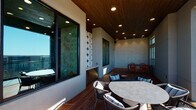 Located at 2405 Marshall St in Fort Worth, TX, this covered outdoor terrace offers a thoughtfully designed gathering space with striking architectural details and panoramic surroundings. The space features rich hardwood decking throughout, providing a warm and cohesive foundation underfoot. A wood-paneled ceiling fitted with recessed lighting overhead creates an inviting, sheltered atmosphere while maintaining an open, airy feel.

Decorative tilework serves as a defining design element, with two distinct patterned accent walls — one featuring a bold geometric circle motif in blue and white, and another with an intricate floral-inspired pattern — adding visual interest and personality to the space. Large floor-to-ceiling windows and glass panel railings allow natural light to flow freely and preserve unobstructed sightlines.

Multiple seating arrangements accommodate a variety of uses. Wicker chairs with navy cushions surround a round table in one area, while vibrant orange chairs paired with a rectangular table occupy a semi-enclosed nook with additional window light. The terrace opens to expansive views of the Fort Worth skyline, the Trinity River, and a pool area below, making it an exceptional vantage point for enjoying the surrounding landscape.