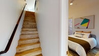 This two-floor space combines contemporary design with warm, functional finishes throughout. Hardwood flooring flows consistently across both levels, connected by a wood-treaded staircase with a clean, modern profile. The open-plan lower level centers around a well-equipped kitchen featuring dark wood cabinetry, a light aqua tile backsplash, stainless steel appliances, and a white quartz peninsula with an undermount sink and bar seating. The adjoining living area is bright and airy, benefiting from a large window with outdoor views of the surrounding neighborhood. The upper level houses the bedroom, a well-proportioned room with multiple windows framing views of an adjacent brick exterior, providing natural light and architectural character. A dedicated closet offers practical storage. Two bathrooms serve the space: one features a glass-enclosed walk-in shower with white square tile surround and terrazzo-style flooring, while the second offers a full soaking tub with matching white tile walls. Both bathrooms are finished with warm wood vanities and stone-toned countertops, maintaining a cohesive, modern aesthetic throughout the home.