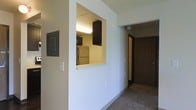 This single-floor residential space is designed with a practical and functional layout. A defining architectural feature is the pass-through window cutout in the wall that connects the kitchen to the adjacent living area, creating an open sightline and enhancing the sense of connectivity between spaces. The kitchen is equipped with dark espresso-toned cabinetry and includes full-size appliances, including a refrigerator, with overhead lighting that keeps the cooking area well-lit. The neutral beige walls and carpeted flooring throughout contribute to a clean, understated aesthetic. A hallway extends from the main living area, providing access to additional rooms via a dark wood-finished door, offering a clear and organized traffic flow through the unit. The ceiling features a textured tile finish, adding subtle dimension overhead. Electrical panel access is integrated discreetly into the main corridor wall, reflecting a thoughtful approach to utility placement. The overall ambiance is modest and straightforward, with a layout that prioritizes efficient use of space and ease of movement between areas. The consistent use of neutral tones and dark wood accents creates a cohesive visual identity throughout the unit.