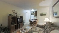 This single-floor space offers a practical and comfortable open-concept layout that seamlessly connects the living room, dining area, and kitchen. The living room is furnished with a patterned sofa and loveseat arranged around a wooden coffee table, complemented by a freestanding entertainment unit housing a television and decorative storage cubbies. Subtle wall décor, including a framed landscape print and a decorative clock, add a homey touch to the neutral-toned walls.

A passage leads directly into the compact dining area, which features a wooden table and matching chairs, providing an intimate space for meals. The kitchen, visible from both the living and dining areas, is equipped with dark wood cabinetry, a white refrigerator, and countertop appliances, offering functional efficiency in a modest footprint.

Carpet flooring runs throughout the living and dining areas, contributing to a warm and quiet ambiance. The layout benefits from a straightforward, accessible flow between rooms, making the space easy to navigate. Overall, this residence presents a well-organized, functional environment suited for comfortable everyday living.
