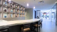 This single-floor space exudes a refined, contemporary aesthetic designed to impress. At its centerpiece is a generously sized curved bar topped with a striking white marble countertop, complemented by dark-stained wood bar seating with tan leather cushions — a combination that balances elegance with comfort. Overhead, sleek pendant lights hang at varying heights, casting a warm, focused glow across the bar surface and enhancing the upscale ambiance.

Along the back wall, a built-in display unit with open cube shelving and integrated recessed lighting provides both functional storage and a sophisticated decorative element, currently showcasing an assortment of decorative bottles. Lower cabinetry with brushed hardware adds to the polished, organized feel of the space.

The interior extends into a bright, open corridor lined with vertical slat partitions that offer a sense of visual depth and separation without fully enclosing the space. Large openings at the far end allow natural light to pour in, connecting the interior to the exterior and giving the space an expansive, welcoming feel. Neutral wall tones, recessed ceiling lighting, and clean tile flooring throughout contribute to the cohesive, modern design. The overall atmosphere is professional, stylish, and thoughtfully curated.