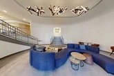 Located at 1027 E Genesee St in Syracuse, New York, this expansive lobby space offers a sophisticated and welcoming environment across a generous single-floor footprint. The centerpiece of the space is a striking rotunda-style lounge area crowned by a coffered oval ceiling recess, from which clusters of sculptural, multi-directional pendant light fixtures hang dramatically overhead. A curved, deep-blue modular sectional sofa anchors the rotunda, complemented by rounded wood-and-metal accent tables, creating an inviting social hub. A grand staircase with arched framing rises prominently from the rear of the rotunda, drawing the eye and adding architectural grandeur. The entrance features a modern revolving glass door alongside traditional double doors, transitioning into the lobby via a carpeted vestibule and large-format light stone tile flooring that extends throughout. Warm wood-paneled accent walls add texture and contrast against the crisp white walls. A secondary lounge area features additional blue seating arrangements, patterned area rugs, and views to the outdoors through large windows. A glass-partitioned private office or consultation room provides functional workspace with natural light. The overall ambiance is polished and contemporary, balancing open communal areas with defined functional zones.