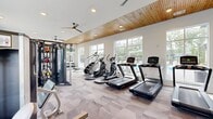 Located at 12801 Lucks Lane in Midlothian, VA, this fitness center offers a generously sized, single-floor layout designed to support a full range of workout routines. The main gym area features a striking combination of a warm natural wood ceiling, crisp white walls, and bold teal accent walls that create an energizing yet welcoming atmosphere. Recessed lighting and dual ceiling fans run the length of the space, ensuring consistent comfort throughout. Large windows line multiple walls, flooding the interior with natural light and providing views of the surrounding outdoor areas, including a glimpse of a pool and landscaped grounds. The main floor is finished with textured patterned carpet and houses an extensive selection of equipment, including treadmills, ellipticals, stair climbers, a multi-function cable machine, selectorized strength machines, a Smith machine with bench, and free weights. A dedicated spin studio adjoins the main floor, finished with light hardwood flooring and wrapped in teal walls. It features cycling bikes, a wall-mounted flat-screen TV with a Fitness on Demand system, a mirrored wall, and a ballet barre — ideal for guided classes. Large sliding glass doors in the spin room open directly to an outdoor area, seamlessly connecting indoor and outdoor fitness experiences.
