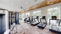 Located at 12801 Lucks Lane in Midlothian, VA, this fitness center offers a generously sized, single-floor layout designed to support a full range of workout routines. The main gym area features a striking combination of a warm natural wood ceiling, crisp white walls, and bold teal accent walls that create an energizing yet welcoming atmosphere. Recessed lighting and dual ceiling fans run the length of the space, ensuring consistent comfort throughout. Large windows line multiple walls, flooding the interior with natural light and providing views of the surrounding outdoor areas, including a glimpse of a pool and landscaped grounds. The main floor is finished with textured patterned carpet and houses an extensive selection of equipment, including treadmills, ellipticals, stair climbers, a multi-function cable machine, selectorized strength machines, a Smith machine with bench, and free weights. A dedicated spin studio adjoins the main floor, finished with light hardwood flooring and wrapped in teal walls. It features cycling bikes, a wall-mounted flat-screen TV with a Fitness on Demand system, a mirrored wall, and a ballet barre — ideal for guided classes. Large sliding glass doors in the spin room open directly to an outdoor area, seamlessly connecting indoor and outdoor fitness experiences.