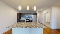 Located at 704 Williamson St in Madison, WI, this well-appointed one-bedroom, one-bathroom unit offers a thoughtfully designed open-concept layout. The combined kitchen and living area serves as the heart of the home, showcasing rich hardwood flooring, crisp white walls, and recessed lighting throughout. The modern kitchen is a standout feature, equipped with dark espresso cabinetry, granite countertops, a large central island with a built-in sink, stainless steel appliances including a refrigerator, range, and microwave, and stylish pendant lighting overhead. The spacious living area flows seamlessly from the kitchen and is bathed in natural light through a bank of large windows offering outdoor views. A wide hallway with recessed lighting connects the living spaces to the private bedroom, which features plush carpeting, a ceiling fan, and windows that allow ample natural light. The bedroom includes access to a generously sized walk-in closet fitted with multi-level wire shelving. The bathroom offers a clean, neutral aesthetic with a full tub-shower combination and tile-style flooring. This unit combines contemporary finishes with a practical, comfortable layout.