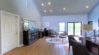 This single-floor space makes a strong first impression with its soaring vaulted ceilings and open, airy layout. The main room is bathed in natural light, drawing from a large glass door that opens to an outdoor deck area surrounded by lush greenery, creating a seamless connection between the interior and exterior environments.

The flooring throughout is warm-toned hardwood, providing a clean and cohesive foundation for the space. Recessed lighting is installed throughout the ceiling, offering ample and well-distributed illumination that complements the natural light.

A central dining area features a round table with modern metal-frame chairs, anchored by a vibrant, patterned area rug that adds a pop of color and visual interest. Along one wall, a dark wood credenza with glass-front cabinet doors serves as both a functional storage unit and a display surface, adorned with decorative items and a coffee station. Adjacent to it, a compact workspace or service counter provides additional utility.

The walls are painted in a soft blue-gray tone, lending the space a calm and welcoming ambiance. Framed artwork adds subtle decorative touches throughout. Overall, the space is thoughtfully arranged to balance functionality and comfort in a bright, well-appointed setting.