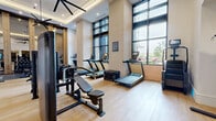 This modern fitness center spans a generous single floor, thoughtfully organized into multiple distinct zones to support a comprehensive workout experience. The main strength and cardio area is the architectural centerpiece, boasting soaring double-height ceilings, a dramatic coffered tray detail, and oversized floor-to-ceiling windows that flood the space with natural light and offer open views to the outdoors. Large-format mirror panels line the walls, visually expanding the room while a statement multi-blade ceiling fan adds both function and design flair. Light hardwood flooring runs throughout, creating a clean, contemporary aesthetic. This primary zone houses a full array of commercial-grade equipment, including treadmills, cable machines, a rowing machine, free weights, and resistance benches, all arranged to maximize usability and flow. Decorative textured wall panels lend warmth and character alongside the exposed light brick accents. Adjacent to the main floor is a dedicated cycling studio, a bright and minimalist room equipped with stationary bikes, a large wall-mounted mirror with an integrated display screen, and recessed lighting. A separate, more intimate training room provides additional space for focused workouts, complete with a smart mirror, connected fitness bike, and built-in cabinetry, all accessible through sleek black-framed glass doors.