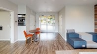 Metro 510's amenity space in Vallejo, California offers a thoughtfully designed single-floor community hub built for social interaction and daily convenience. The entry lobby sets a clean, modern tone with wood-look flooring, glass double doors that invite natural light, and casual seating including a compact round table with vibrant orange chairs and a contemporary sofa — ideal for informal meetings or relaxed waiting.

The centerpiece of the space is a well-appointed kitchen and dining area. Dark espresso cabinetry with brushed nickel hardware lines the walls, complemented by a crisp white subway tile backsplash and light quartz countertops. A deep stainless steel sink with a pull-down faucet serves the functional prep area, while a coffee and beverage station provides convenient refreshment options. A large natural wood communal dining table surrounded by modern white shell chairs accommodates a generous number of residents at once.

A striking reclaimed wood accent wall serves as the backdrop for the branded Metro 510 lounge area, where modular blue-and-gray seating, a marble-top coffee table, and warm accent pieces create an inviting retreat. Additional café-style seating along another wood-paneled wall offers flexible dining arrangements. Throughout, wood-look flooring, neutral walls, and coordinated pops of orange and blue give the space a cohesive, contemporary aesthetic.