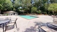 Located at 7403 Treehills Pkwy in Stone Mountain, Georgia, this outdoor amenity space centers around a generously sized swimming pool with clear turquoise water, framed by a red-trimmed pool edge and a wide concrete sundeck. The pool area is designed for both relaxation and recreation, offering ample deck space for lounging and movement around the water.

A dedicated seating area lines the perimeter, furnished with multiple sling-style lounge chairs and standard patio chairs positioned to take advantage of the surrounding natural scenery. The space is enclosed by a classic black wrought iron fence, providing security while maintaining an open, airy feel. Decorative lamp posts accent the fencing, adding a polished, community-oriented aesthetic.

The backdrop is one of the space's most notable features — mature Georgia pines and a dense canopy of deciduous trees surround the area on all sides, creating a shaded, park-like atmosphere that adds privacy and natural beauty. A stone retaining wall at the far end of the pool adds architectural character, while a small utility or restroom structure is visible nearby. The overall ambiance is tranquil and well-maintained, offering a pleasant outdoor retreat within a residential community setting.