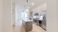 This contemporary single-floor residence showcases a thoughtfully designed open-concept layout that seamlessly connects the kitchen and living areas. The kitchen serves as the centerpiece of the home, featuring a generous island with a crisp white quartz countertop and a gray base, illuminated by a pair of stylish pendant lights. Two-tone cabinetry — combining light wood upper cabinets with glass-front accents and gray lower cabinets — frames a full suite of stainless steel appliances, including a gas range, built-in microwave, refrigerator with ice maker, and dishwasher. A white subway tile backsplash adds a clean, classic touch throughout the cooking space. Light-toned hardwood flooring runs consistently throughout the residence, contributing to the bright, airy ambiance. The bedroom is a serene retreat, bathed in natural light through large windows with views to the surrounding neighborhood, and includes a spacious walk-in closet outfitted with dual hanging rods and shelving. The bathroom is elegantly appointed with large-format marble tile surrounding a soaking tub and separate glass-enclosed shower, complemented by a dual-sink vanity with white countertops, dark hardware accents, and a wide frameless mirror — offering a refined, spa-like experience.