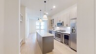 This contemporary single-floor residence showcases a thoughtfully designed open-concept layout that seamlessly connects the kitchen and living areas. The kitchen serves as the centerpiece of the home, featuring a generous island with a crisp white quartz countertop and a gray base, illuminated by a pair of stylish pendant lights. Two-tone cabinetry — combining light wood upper cabinets with glass-front accents and gray lower cabinets — frames a full suite of stainless steel appliances, including a gas range, built-in microwave, refrigerator with ice maker, and dishwasher. A white subway tile backsplash adds a clean, classic touch throughout the cooking space. Light-toned hardwood flooring runs consistently throughout the residence, contributing to the bright, airy ambiance. The bedroom is a serene retreat, bathed in natural light through large windows with views to the surrounding neighborhood, and includes a spacious walk-in closet outfitted with dual hanging rods and shelving. The bathroom is elegantly appointed with large-format marble tile surrounding a soaking tub and separate glass-enclosed shower, complemented by a dual-sink vanity with white countertops, dark hardware accents, and a wide frameless mirror — offering a refined, spa-like experience.