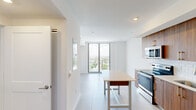 Located on the 15th floor at 218 NW Eighth St in Miami's vibrant urban core, this modern residence offers a bright, open-concept layout finished throughout with large-format porcelain tile flooring. The living and dining area flows seamlessly from the kitchen toward expansive floor-to-ceiling windows and a private balcony, framing sweeping views of the Miami skyline and surrounding greenery. Natural light fills the interior throughout the day, accentuating the clean white walls and contemporary design.

The kitchen is a standout feature, outfitted with rich walnut-toned cabinetry, gold-accented hardware, a white subway tile backsplash, and a full suite of stainless steel appliances including a French door refrigerator, electric range, built-in microwave, and dishwasher. A central island provides additional prep and dining space.

The bedroom is generously sized, featuring a ceiling fan, a wall of closet space, and its own floor-to-ceiling window with continued city views. The bathroom is elegantly appointed with marble-look tile surrounding a soaking tub and shower combination, a warm wood vanity, white countertop, and a globe pendant light fixture. A secondary room offers flexible use as a bedroom, office, or den. The unit is accessed through a bold navy blue entry door.