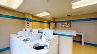 Located at 150 Arbors Cir in Gahanna, Ohio, this shared laundry facility is a practical and well-maintained amenity designed to serve its community. The room is finished with warm yellow walls accented by a bold teal horizontal stripe, giving the space a clean and recognizable character. Overhead, recessed fluorescent lighting panels provide bright, even illumination throughout the area, ensuring visibility and ease of use at any time of day.

A row of top-loading washing machines lines the main wall, each equipped with clearly posted operating instructions for user convenience. A step-by-step guide is also displayed near the entrance, walking users through the process of operating the machines. Adjacent to the washers, a smooth countertop surface offers space for folding or sorting laundry.

A community bulletin board mounted on the wall serves as an informational hub, displaying notices and resources for residents. Safety is also addressed with a fire extinguisher mounted and readily accessible on the wall. The flooring is durable and easy to clean, suited for the high-traffic demands of a shared utility space. Overhead ventilation ensures the room remains comfortable during use. Overall, this laundry facility is a functional, clearly organized shared amenity designed with everyday usability in mind.