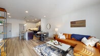 This well-appointed single-floor residence showcases a thoughtfully designed open-concept layout that seamlessly connects the living, dining, and kitchen areas. Light-toned wood-style flooring runs throughout, complementing the clean white walls and recessed lighting that keep the space feeling bright and airy.

The living area is anchored by large windows and a private exterior door, allowing generous natural light to fill the room. The dining area sits adjacent, centered around a round dark-wood table beneath a round accent mirror, creating an intimate yet open atmosphere.

The kitchen is a standout feature, boasting two-tone cabinetry with crisp white upper cabinets and rich dark lower cabinets, granite countertops, stainless steel appliances, and a peninsula with bar seating. Under-cabinet lighting adds both function and ambiance. A pantry is conveniently accessible directly from the kitchen.

The bedroom is serene and well-lit, featuring a large window that invites natural light, with ample space for a full-sized bed and furnishings. The bathroom includes a walk-in glass-enclosed shower with matte black fixtures, a rainfall showerhead, and a clean, minimalist aesthetic.

A dedicated in-unit laundry area with full-sized washer and dryer units rounds out this practical and polished residence.