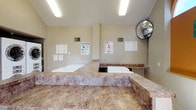 This shared laundry facility is a single-floor space designed with practicality and functionality in mind. The room features vaulted ceilings that lend a sense of openness, while large windows along one wall allow natural light to filter in, offering views of surrounding greenery. Light tile flooring runs throughout, providing a clean and easy-to-maintain surface.

The facility is equipped with multiple stacked and front-loading washers and dryers, arranged along the walls to maximize usable floor space. Generous granite-style laminate folding countertops extend along the perimeter of the room, offering ample surface area for sorting and folding laundry. A dedicated utility sink with matching granite-style surround provides added convenience for hand-washing or pre-treating garments.

Informational signage, including laundry tips and usage instructions in multiple languages, is posted throughout the room, reflecting its communal nature. A convex safety mirror is mounted in a corner for enhanced visibility. Overhead fluorescent lighting ensures the space remains well-lit at all times. A small seating stool near the window area offers a comfortable spot for waiting. The overall ambiance is clean, organized, and user-friendly, making it a well-considered shared amenity.