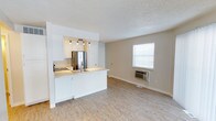 This single-floor space features an open-concept design that seamlessly connects the kitchen and living area, creating a bright and functional layout. The kitchen is outfitted with white cabinetry, light-toned countertops, a stainless steel refrigerator, and a built-in cooktop with a sink along the peninsula-style counter. Track lighting above the kitchen provides focused illumination, while the white cabinetry and clean finishes contribute to a fresh, modern aesthetic. The peninsula serves as a natural divider between the kitchen and the open living area without sacrificing the sense of spaciousness. The living area is finished with light hardwood-style flooring that extends throughout, adding warmth and continuity to the space. A window fitted with white horizontal blinds allows natural light to filter in, brightening the neutral-toned walls. A wall-mounted air conditioning unit is positioned beneath the window, providing climate control. The textured ceiling is consistent throughout, and white baseboards frame the perimeter of the space. Overall, the layout is practical and well-lit, with a clean, minimalist character suited to a variety of uses.