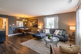 This two-floor residence offers a thoughtfully designed layout with modern finishes and a cohesive color palette throughout. The open-concept living and dining area anchors the main floor, featuring rich hardwood-style flooring, neutral-toned walls, and an abundance of natural light streaming through multiple windows dressed with blinds and sheer curtains. The living room comfortably accommodates a large sectional sofa and a glass coffee table set atop a geometric-patterned area rug, while a corner provides additional seating. Adjacent to the living space, the dining area is highlighted by a chandelier overhead and a pass-through window connecting directly to the kitchen, creating a seamless flow between spaces. The kitchen is equipped with stainless steel appliances, including a refrigerator, range, and microwave, complemented by white cabinetry and ample counter space. The bedroom is a standout space, featuring a bold teal accent wall, a ceiling fan, closet storage, and a sun-filled window. The bathrooms include a full bath with over-toilet cabinetry and a framed mirror, as well as a half bath. Dark hardwood-style flooring runs consistently through the space, tying together the home's warm and contemporary aesthetic.