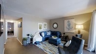 This single-floor residence offers a comfortable and thoughtfully designed living space that blends modern style with a relaxed, coastal-inspired aesthetic. The living room serves as the heart of the home, showcasing warm neutral wall tones complemented by rich hardwood-style flooring that grounds the space with a sense of warmth. A textured ceiling adds architectural character overhead. The room is anchored by a bold navy blue sectional sofa paired with a matching ottoman, creating an inviting and cohesive seating arrangement. A geometric-patterned area rug in shades of blue, gold, and green adds visual interest underfoot. Wall décor includes botanical prints and a coral-themed artwork, reinforcing the coastal theme, while a round decorative mirror introduces an elegant focal point. Accent side tables with slender metallic frames provide functional surfaces without overwhelming the layout. Potted greenery placed throughout the room brings a touch of nature indoors and enhances the overall freshness of the design. The space is well-proportioned, offering a natural flow and a bright, airy ambiance that makes it both functional and welcoming for everyday living and entertaining alike.