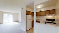 Located at 275 Glen Riddle Road in Media, Pennsylvania, this single-floor residence offers a clean, functional layout designed for comfortable everyday living. The kitchen is a practical and well-equipped space, featuring warm-toned wooden cabinetry that provides ample storage, a white refrigerator, a built-in dishwasher, and a sink set beneath upper cabinets. Light-toned tile flooring runs throughout the kitchen, contributing to a bright and easy-to-maintain environment. A structural dividing wall separates the kitchen from the adjoining living area, creating a sense of definition between spaces while maintaining an open feel. The living area is finished with plush wall-to-wall carpeting in a neutral tone, complementing the crisp white walls and contributing to a warm, inviting atmosphere. Natural light filters into the living space through a window, enhancing the airy quality of the room. The overall palette of the residence — soft whites, warm wood tones, and neutral textiles — creates a cohesive and understated aesthetic. The single-floor layout ensures easy accessibility and a seamless flow throughout the home, making it a straightforward and welcoming space in the Media, PA area.