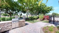 This single-story property showcases a thoughtfully designed outdoor living area as a standout feature. The spacious paver patio is finished in a natural stone pattern, providing a durable and attractive surface for outdoor entertaining. At its centerpiece sits a built-in outdoor kitchen station, constructed with stacked stone cladding and equipped with a stainless steel grill and cabinetry, offering both functionality and aesthetic appeal. Matching stone columns frame the space, adding architectural structure and definition to the patio area.

The surrounding landscape is lush and well-maintained, featuring mature shade trees that provide generous canopy cover, along with tropical plantings including ornamental grasses and colorful foliage that enhance the tropical character of the setting. Beyond the landscaping, a serene water feature is visible in the background, adding a tranquil element to the outdoor environment. The overall ambiance of the exterior is relaxed and resort-like, ideal for outdoor gatherings. The remaining interior images were too underexposed to provide detailed descriptions of the indoor spaces.
