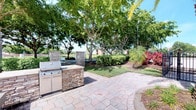 This single-story property showcases a thoughtfully designed outdoor living area as a standout feature. The spacious paver patio is finished in a natural stone pattern, providing a durable and attractive surface for outdoor entertaining. At its centerpiece sits a built-in outdoor kitchen station, constructed with stacked stone cladding and equipped with a stainless steel grill and cabinetry, offering both functionality and aesthetic appeal. Matching stone columns frame the space, adding architectural structure and definition to the patio area.

The surrounding landscape is lush and well-maintained, featuring mature shade trees that provide generous canopy cover, along with tropical plantings including ornamental grasses and colorful foliage that enhance the tropical character of the setting. Beyond the landscaping, a serene water feature is visible in the background, adding a tranquil element to the outdoor environment. The overall ambiance of the exterior is relaxed and resort-like, ideal for outdoor gatherings. The remaining interior images were too underexposed to provide detailed descriptions of the indoor spaces.