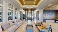 Located on Sydney Road in Morganville, NJ, this single-floor community amenity space offers a thoughtfully designed and generously proportioned environment spanning approximately 3,000 square feet. The grand main living area immediately impresses with soaring tray ceilings finished in gold tones, recessed lighting, and a striking geometric chandelier. Polished tile flooring, a decorative lattice room divider, and carefully curated furnishings — including neutral sofas and warm leather accent chairs — create an inviting, upscale atmosphere. A second living area provides a more relaxed lounge setting, featuring built-in patterned bench seating, sculptural wall art, modular soft seating, and a wall-mounted television, bathed in natural light from large windows. The dining area offers a bright, casual setting with high-top seating and generous window views of the exterior grounds. The well-appointed exercise room is a standout feature, housing a full array of cardio machines including treadmills and ellipticals, strength training equipment, weight benches, and a multi-station cable machine. Large windows and wall mirrors amplify the sense of space and allow natural light to flood in, complemented by a ceiling fan overhead. A connecting hallway ties all areas together seamlessly, completing this refined and functional community facility.