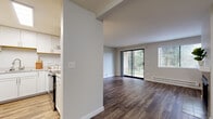 This single-floor residence is designed with a clean, modern aesthetic and a practical open layout. Wood-style flooring runs throughout the main living areas, lending warmth and continuity to the space. The living room serves as the central gathering area, highlighted by a tile-surround fireplace and generous natural light streaming in through a large sliding glass door and an additional window fitted with blinds. The sliding door opens to an outdoor view of mature trees, creating a pleasant connection to nature.

The kitchen is thoughtfully designed with white cabinetry, a stainless steel refrigerator, a stainless range, a wall-mounted range hood, and light countertops. A half-wall peninsula separates the kitchen from the main living area, providing both visual openness and functional counter space. A skylight above the kitchen enhances brightness in the workspace. Baseboard heating units are present along the walls throughout the unit.

The bedroom features plush carpeting, a natural window with blinds, and a set of sliding closet doors with built-in shelving and hanging rods, offering ample storage. The bathroom includes a full-sized soaking tub with a shower surround and chrome fixtures. The entry area is defined by a paneled white door and smooth neutral walls, providing a tidy transition into the home.