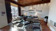 Located at 12103 Inwood Circle in Dallas, Texas, this single-story sales office delivers a compelling blend of Texas heritage aesthetic and contemporary design. The space makes an immediate impression at entry, where rich wide-plank hardwood flooring and dramatic black steel-framed glass partitions define the layout with industrial precision. A striking arched passageway with matching black steel detailing serves as a prominent architectural focal point, guiding visitors deeper into the interior.

The main workspace is anchored by a bold white brick accent wall bearing the "Republic of Texas Pioneers" mural — a nod to Texas history that sets a distinctly regional tone. Warm wood-paneled ceilings contrast beautifully against the white walls, while oversized black lantern-style chandeliers provide ambient and accent lighting throughout. A sleek linear fireplace adds an inviting warmth to the consultation area, which is furnished with contemporary desks and modern lounge seating arranged to facilitate client interaction.

Large steel-framed windows flood the space with abundant natural light, offering views to the exterior streetscape. Additional private offices and corridors branch off the main area, separated by the signature glass partition walls that maintain visual openness while providing functional division. The overall ambiance is professional yet warmly approachable, reflecting a refined Texas character throughout.