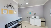 This single-story laundry facility in Lewisville is designed with functionality and convenience in mind. The room features light gray walls accented by white crown molding and baseboards, creating a clean, neutral atmosphere. Recessed ceiling lighting provides ample illumination throughout the space. Two white top-loading washing machines are positioned along the back wall, equipped with coin-operated controls for shared use. A wall-mounted clothes rod offers a convenient spot for hanging garments, while a white folding table provides a dedicated surface for sorting and folding laundry. A black metal bench with a lattice-style seat is available for residents waiting during their laundry cycle. Decorative laundry-themed signs add a touch of personality to the walls, and a small bulletin board area near the entrance allows for posted notices or announcements. The light-toned speckled flooring is easy to maintain and well-suited for a high-traffic utility space. Overall, the room is thoughtfully arranged to accommodate multiple users comfortably while maintaining an organized and welcoming environment.