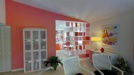 This single-floor space makes a bold visual statement through its use of warm coral and pink tones across the walls, creating an energetic and inviting atmosphere throughout. A standout architectural feature is the open-frame room divider, composed of suspended rectangular panels in varying shades of pink, coral, and white. This eye-catching element serves as both a visual focal point and a subtle partition between the living and dining areas, allowing light to pass freely while defining each zone with style.

The living area features white lattice-back chairs that complement the crisp white trim and furnishings found throughout the space. A white mesh-front storage cabinet along one wall adds both function and texture. A lush green potted plant introduces a refreshing natural contrast to the warm color palette.

Beyond the divider, the dining area is furnished with a round white table and matching chairs, maintaining the clean, cohesive aesthetic. Recessed lighting in the ceiling and a modern table lamp provide layered illumination. Colorful wall art adds an additional layer of personality. Overall, the space balances playful design with practical layout and a cohesive, contemporary aesthetic.