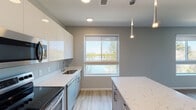 This single-floor space showcases a thoughtfully designed modern interior with a cohesive neutral palette and contemporary finishes throughout. The kitchen serves as a standout feature, equipped with high-gloss white upper cabinetry, wood-toned lower cabinets, and a striking quartz island with veined marble-like detailing. Stainless steel appliances, including a range, microwave, and dishwasher, complete the culinary space. Large windows frame serene waterfront views, flooding the kitchen with natural light, while sleek pendant lights add a refined accent overhead.

The kitchen flows seamlessly into an open living and dining area, where light wood-look flooring continues underfoot. Multiple windows and a sliding glass door provide access to a balcony, further enhancing the connection to the outdoor waterfront scenery. The living area offers a clean, versatile layout with neutral walls.

Bedrooms feature plush carpeting and generously sized windows with views of surrounding greenery, creating calm, comfortable retreats. The bathroom is finished with large-format tile flooring, a wood-toned vanity with an extended countertop, and a wide mirror, offering both style and functionality.