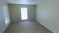 This single-floor space presents a straightforward and functional layout designed for comfortable everyday living. The interior is finished with neutral, warm beige walls that create a calm and understated atmosphere throughout. The flooring consists of wall-to-wall carpet in a complementary tan tone, adding a layer of softness underfoot and contributing to the overall warmth of the space. A textured popcorn ceiling runs overhead, a classic finish that adds subtle visual interest while maintaining a simple, no-frills aesthetic. Natural light is introduced through a window fitted with horizontal blinds, allowing for adjustable light control while keeping the room bright and airy. Clean white baseboards frame the perimeter of the room, providing a crisp contrast against the beige walls and carpet. The space is open and uncluttered, offering a versatile blank canvas suitable for a variety of uses and furniture arrangements. Electrical outlets are positioned along the walls, ensuring practical functionality throughout. Overall, the space conveys a modest, move-in-ready quality with its neutral palette, soft flooring, and ample natural light.
