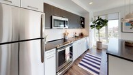 This 1-bedroom, 1-bathroom residence is designed with a clean, contemporary aesthetic throughout. The living area is anchored by large floor-to-ceiling windows on multiple walls, flooding the space with natural light and offering sweeping elevated views of the surrounding cityscape and greenery. The open layout connects the living area to the kitchen seamlessly, with hardwood-style flooring running throughout. A dedicated media wall provides a functional focal point, while the versatile layout accommodates both lounging and dining comfortably.

The kitchen features a galley-style design with stainless steel appliances, including a refrigerator, electric range, built-in microwave, and dishwasher. Dark upper cabinetry contrasts with white lower cabinets and a white tile backsplash, while dark countertops complete the modern palette. A window at the end of the kitchen brings in additional natural light.

The bedroom is generously proportioned, with a large picture window framing elevated outdoor views. Warm wood tones from matching furniture pieces complement the neutral walls and plush area rug.

The bathroom maintains the unit's modern character with a floating white vanity, illuminated mirror, tile flooring, and clean white finishes throughout.