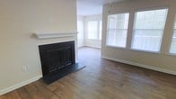 This single-floor space offers a welcoming living area centered around a traditional fireplace with a crisp white mantel and black surround, serving as a natural focal point. Rich, dark hardwood flooring extends throughout, adding warmth and character to the interior. A textured ceiling adds subtle architectural detail overhead.

Natural light flows generously into the space through multiple windows fitted with white horizontal blinds, including a notable bay-style window arrangement in the corner that brightens the room and creates an open, airy atmosphere. The neutral, light-toned walls complement the darker flooring, creating a balanced contrast throughout.

The open layout of the living area allows for flexible furniture arrangement, making it well-suited for both everyday living and entertaining. Clean baseboards and understated finishes contribute to a tidy, move-in-ready aesthetic. The overall ambiance is calm and functional, blending classic design elements with practical, comfortable living — ideal for those seeking a straightforward, well-appointed space.