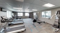 This fitness center offers a comprehensive workout environment designed to accommodate a wide range of exercise routines. The space features a drop-panel ceiling with recessed fluorescent lighting that ensures the room is consistently and evenly illuminated. The flooring consists of patterned low-pile carpet with teal accent stripes, providing both comfort and durability underfoot.

The cardio section is well-appointed with multiple treadmills and elliptical machines, each positioned within easy view of wall-mounted flat-screen televisions. A rowing machine further expands the cardio options available to users. For strength training, the space includes multiple cable and pulley machine systems, a multi-function weight machine, a free weight rack stocked with a full range of dumbbells, and an adjustable workout bench. Exercise balls and a foldable fitness mat round out the functional training equipment.

Windows along the perimeter walls allow natural light to filter in and offer views of the surrounding outdoor areas, including a glimpse of an adjacent pool area visible through interior windows. A large wall mirror enhances the sense of openness while allowing users to monitor their form. The overall atmosphere is clean, functional, and well-organized, making it a practical and motivating environment for daily fitness activities.