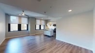 This single-floor unit showcases a thoughtfully designed open-concept layout that seamlessly integrates the living and kitchen areas into one cohesive, light-filled space. Light-toned hardwood flooring runs throughout, lending warmth and continuity to the interior.

The kitchen is a standout feature, outfitted with sleek flat-front cabinetry in a soft white finish, dark granite countertops, and a full suite of stainless steel appliances including a refrigerator, range, built-in microwave, and dishwasher. A mobile island with a dark countertop provides additional workspace and flexibility, while a glass pendant light adds a refined touch above. A white tile backsplash completes the clean, modern aesthetic.

The open living area is generously proportioned, bathed in natural light from multiple windows dressed with neutral roller shades. A contemporary ceiling fan with a brushed gold finish serves as a subtle focal point overhead, while recessed lighting ensures the space is well-lit throughout.

The bathroom is clean and minimal in design, featuring white fixtures, a wall-mounted towel bar, and a large mirror with integrated lighting, contributing to a bright and functional atmosphere.

Overall, the space presents a modern, move-in ready environment with a neutral palette, quality finishes, and a practical open floor plan well-suited for everyday living.