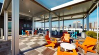 This rooftop space combines covered and open-air areas to create a dynamic, multi-functional outdoor environment high above the surrounding cityscape. The open-air deck features a wood-tiled floor laid in a checkerboard pattern, furnished with bold orange, yellow, teal, and green Adirondack-style chairs and low round tables that encourage casual gathering. A partially covered pavilion section extends the usable space, supported by white steel columns and fitted with a paneled ceiling that houses overhead heaters and recessed lighting, allowing for year-round use. Open on all sides, the pavilion captures sweeping panoramic views of the urban skyline and horizon beyond. Additional dining-height tables and benches are positioned within the covered zone, offering a more structured seating arrangement. The interior entry area features vibrant teal walls, large floor-to-ceiling windows that flood the space with natural light, elevator access, and a stairwell entry door, making the rooftop easily accessible. The combination of lively furnishings, generous natural light, open sightlines, and city views gives this rooftop an energetic yet comfortable ambiance suited for both socializing and quiet relaxation.