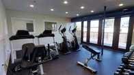 This fitness center offers a clean, functional environment designed to support a well-rounded workout routine. The space is outfitted with a selection of cardio equipment, including treadmills and elliptical machines, positioned to take full advantage of the natural light that pours in through large floor-to-ceiling windows and glass doors along one wall. These expansive windows provide views of an outdoor deck area and surrounding greenery, creating an open, airy atmosphere that makes exercising more enjoyable. A weight bench and additional strength training equipment round out the available amenities, catering to a variety of fitness needs. The flooring is finished in durable dark rubber matting, practical for high-traffic workout use and easy maintenance. Recessed lighting is installed throughout the ceiling, ensuring the space remains well-lit during any hour of the day. A wall-mounted mirror near the entrance adds to the sense of openness and allows users to monitor their form. The light-colored walls and white trim contribute to a bright, inviting ambiance. An emergency exit is clearly marked, and the layout allows for comfortable movement between stations, making this a practical and welcoming space for daily fitness activity.