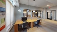This thoughtfully designed single-floor professional space blends functionality with modern aesthetics to create a productive and welcoming work environment. The open-plan layout accommodates a shared workstation area furnished with sleek wooden desks, ergonomic seating, and polished pendant lighting that hangs from elevated ceilings, lending the space an airy, contemporary feel.

Glass-enclosed conference rooms with black-framed partitions provide semi-private meeting spaces while maintaining visual connectivity throughout the office. These rooms are distinguished by their accent walls of reclaimed wood paneling, adding a warm, textural contrast to the otherwise clean, neutral palette of gray walls and carpeted flooring.

A comfortable lounge nook near floor-to-ceiling windows offers an abundance of natural light and exterior views, furnished with a cushioned armchair and side tables — ideal for informal meetings or quiet work. A credenza area along one wall provides additional workspace and storage, accented by a vibrant piece of abstract artwork that adds a splash of color to the space.

Throughout, the design maintains a cohesive, professional atmosphere with neutral tones, recessed lighting, and carefully curated furnishings. The combination of open collaboration areas and enclosed meeting rooms makes this space well-suited for a variety of professional needs.