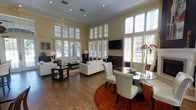 This single-floor space makes a strong architectural impression with its dramatically tall ceilings and an expansive wall of oversized windows fitted with plantation shutters and sheer curtains. The generous fenestration floods the interior with natural light, creating a bright and airy atmosphere throughout.

The open-plan layout seamlessly connects the living and dining areas. The living space is anchored by white upholstered sofas arranged around a dark wood coffee table, all set atop a warm-toned area rug over rich hardwood flooring. A wall-mounted flat-screen television is positioned above a fireplace, serving as a focal point for the seating arrangement. Recessed and track lighting complement the natural light overhead.

The dining area features a round glass-top table surrounded by white upholstered chairs with dark wood frames, set on a deep rust-colored circular rug that contrasts elegantly with the hardwood floor. Decorative floral accents and carefully selected artwork add refined finishing touches. The overall ambiance is contemporary and sophisticated, with a neutral warm color palette that ties the open living and dining zones together cohesively.