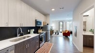 Located at 3200 Dr. Andrew J. Brown Ave in Indianapolis, IN, this thoughtfully designed single-floor unit offers a contemporary living experience within a compact, well-utilized footprint. The open-concept layout seamlessly connects the kitchen and living area, creating a cohesive and functional space.

The kitchen is outfitted with modern two-tone cabinetry — light upper cabinets paired with dark lower cabinets — complemented by a sleek white countertop, a dark tile backsplash, and stainless steel appliances including a refrigerator, gas range, and built-in microwave. The living area is bright and welcoming, benefiting from natural light delivered through a glass exterior door and a large window, both fitted with blinds and curtains. Durable wood-look flooring runs throughout, tying the spaces together cohesively.

The unit includes two bathrooms, one of which features a dark wood vanity with a white countertop, a well-lit mirror, and a full bathtub with shower. A spacious walk-in closet with built-in hanging rods and wood-look flooring provides ample storage. The clean, neutral color palette and modern finishes throughout give the space a fresh, contemporary feel ideal for comfortable everyday living.