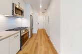 This single-floor unit offers a thoughtfully designed layout with clean, modern aesthetics evident throughout. The open-plan kitchen and living area forms the heart of the space, where warm hardwood-style flooring flows continuously underfoot, complemented by crisp white walls and recessed lighting overhead. The kitchen is equipped with stainless steel appliances, including a gas range, refrigerator, and over-range microwave, set against white cabinetry and a classic subway tile backsplash. A hallway with panel-style doors featuring matte black hardware connects the main living areas, maintaining a cohesive contemporary design language. The living room benefits from natural light streaming through a pair of windows, with a ceiling fan overhead providing airflow and comfort. A standout design element is a bold, textured concrete accent wall that adds an industrial character to the space. The bathroom is well-appointed with large-format tile flooring, a full-width white vanity with under-mount sink, matte black fixtures and hardware, ample cabinetry for storage, and a glass-enclosed walk-in shower featuring striking marble-style tile from floor to ceiling. Overall, the unit presents a cohesive modern design with a neutral palette and quality finishes throughout.