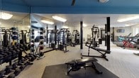 This fitness center is thoughtfully designed to accommodate a wide variety of workout styles and fitness goals. The main exercise floor is expansive and open, featuring light-toned carpet flooring, bold dark structural columns, and a striking teal-blue ceiling accent that adds a modern, energetic atmosphere. Large wall-to-wall mirrors line portions of the space, enhancing the sense of openness and allowing users to monitor their form throughout their workout.

The strength training area is well-appointed with multiple cable machines, weight racks stocked with dumbbells, barbells, and medicine balls, as well as adjustable benches. A dedicated cardio section houses treadmills, upright and recumbent bikes, and ellipticals, with wall-mounted televisions nearby for entertainment. Sanitation stations are conveniently placed throughout the floor. Ceiling fans provide additional air circulation across the space.

A separate studio room offers a quieter environment for flexibility and mind-body training, equipped with yoga mats, stability balls, BOSU domes, and hardwood-style flooring. Natural light fills the studio through large windows. Additional amenities include a tanning booth and a vending machine room accessible via a side corridor, rounding out this comprehensive and well-maintained fitness facility.