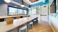 Located at 711–755 Fogg St in Nashville, TN, this well-appointed amenity lounge offers a generously sized, single-floor layout designed to serve as a social and recreational hub. The space is immediately distinguished by its bold design choices, including vivid blue accent walls adorned with large-scale tropical leaf wallpaper in gold and white tones, creating a resort-inspired atmosphere throughout.

Light wood flooring runs continuously across the open floor plan, unifying the various functional zones within the space. A floor-to-ceiling glass curtain wall along one side floods the interior with abundant natural light and frames sweeping views of the Nashville skyline, with direct access to an outdoor terrace featuring a pool area visible beyond.

A lengthy terrazzo-topped bar counter anchors one section of the space, lined with contemporary stools and illuminated by statement pendant lighting. Adjacent to it, a built-in banquette seating area with dining tables provides comfortable gathering spots beneath framed artwork. A slatted wood ceiling detail with integrated linear lighting adds architectural interest in the lounge zone, where a ping-pong table and casual seating arrangements with woven accent chairs complete the amenity-rich environment. The overall ambiance is lively, modern, and thoughtfully designed for social engagement.