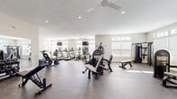 Located in Reynoldsburg, Ohio, this fitness center offers a spacious, single-floor layout designed to accommodate a comprehensive range of workout activities. The interior is finished with neutral walls and durable speckled rubber flooring, creating a clean and functional environment well-suited for exercise.

The cardio zone features multiple treadmills and elliptical machines arranged along a mirrored wall, with wall-mounted flat-screen TVs positioned for convenient viewing during workouts. Ceiling fans provide air circulation throughout the space, enhancing comfort during use.

The strength training area is equipped with a multi-station cable machine, a StairMaster, and various selectorized resistance machines, offering options for both upper and lower body conditioning. A dedicated free weights section includes a full dumbbell rack paired with an adjustable weight bench and a large wall mirror, supporting a variety of strength and toning exercises.

Natural light enters generously through rows of windows positioned along multiple walls, brightening the entire floor. Recessed lighting supplements the natural light for consistent illumination. A separate alcove area provides space for functional training accessories, including stability balls and additional equipment. The fitness center connects to an adjacent room visible through glass-paneled doors, extending the overall usable area of the facility.