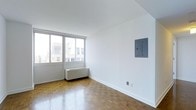 This single-floor space presents a clean, minimalist aesthetic defined by crisp white walls and warm parquet wood flooring laid in a traditional herringbone-style pattern, adding a timeless character to the interior. The main living area is bright and open, benefiting from a large, multi-pane window that spans a generous portion of the exterior wall, allowing abundant natural light to fill the room while offering views of the surrounding urban streetscape. A baseboard heating unit is positioned beneath the window, providing efficient climate control. A flush-mounted electrical panel on the adjacent wall offers a practical, unobtrusive utility feature. The neutral color palette and simple finishes throughout create a versatile backdrop well-suited for a variety of interior arrangements. The overall ambiance is airy and understated, with the natural warmth of the parquet floors providing a pleasing contrast to the bright white walls and ceiling, giving the space a balanced and inviting character.